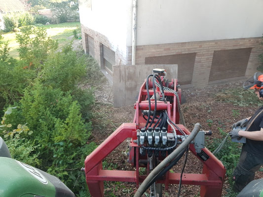 Wurzelfräse arbeitet in enger Gartenecke hinter dem Haus, angrenzende Bepflanzung wird geschont.